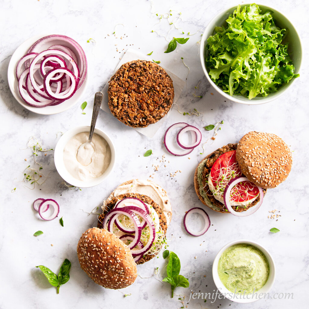 Walnut Pesto Burgers Sandwiches in the making with sprouts, vegan mayo, red onion, tomato, pesto, and a bowl of lettuce.