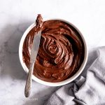 Luscious sugar-free carob frosting in a white bowl with some pulled out on a knife and a towel beside.