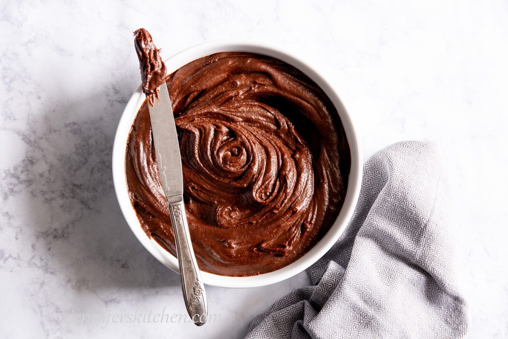 Luscious sugar-free carob frosting in a white bowl with some pulled out on a knife and a towel beside.
