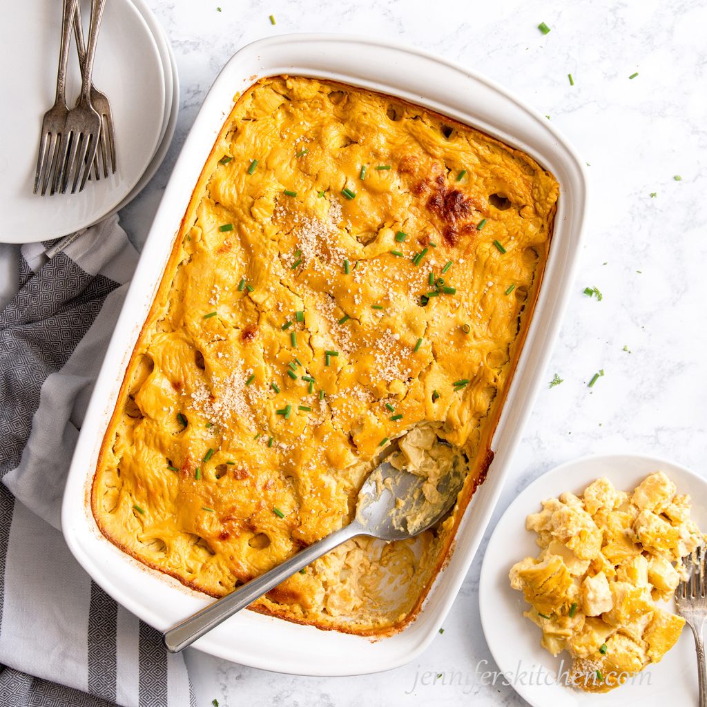 Cheesy Potato Casserole in a white dish topped with fresh chives and vegan parmesan with a plate served out on the side.