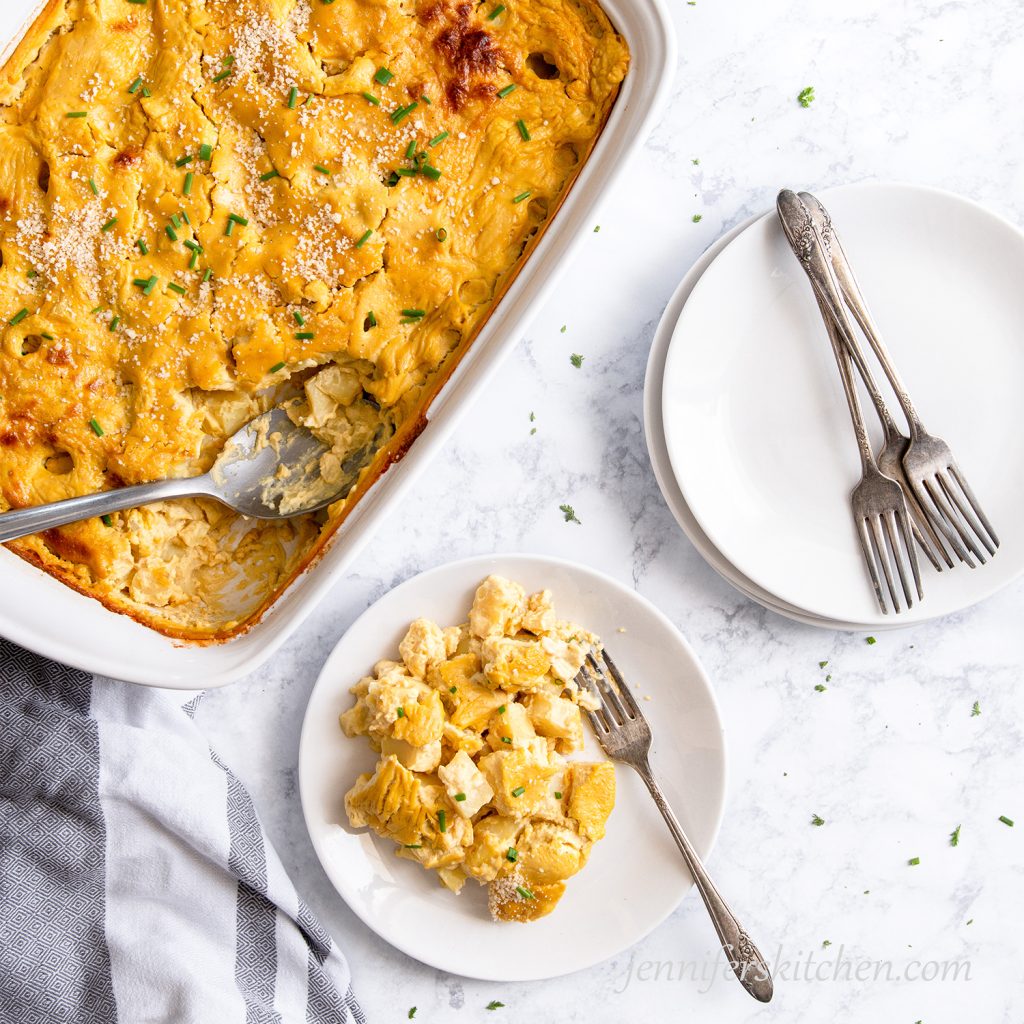 Cheesy Potato Casserole served out on a plate topped with fresh chives and vegan parmesan with a white casserole dish on the side.