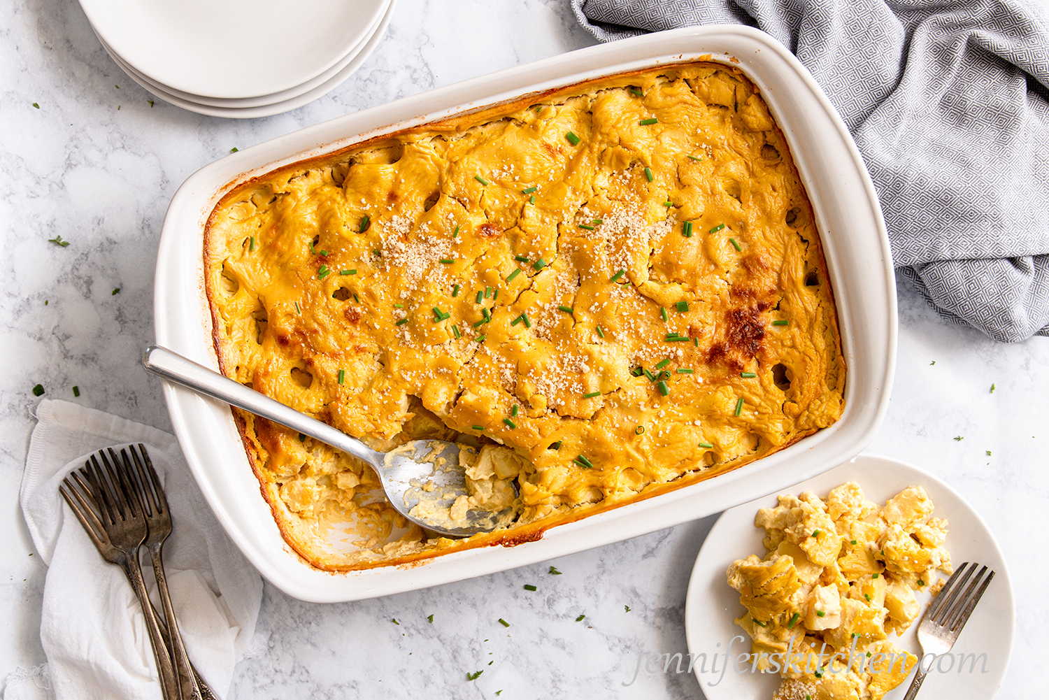 Cheesy Potato Casserole in a white dish topped with fresh chives and vegan parmesan with a plate served out on the side.