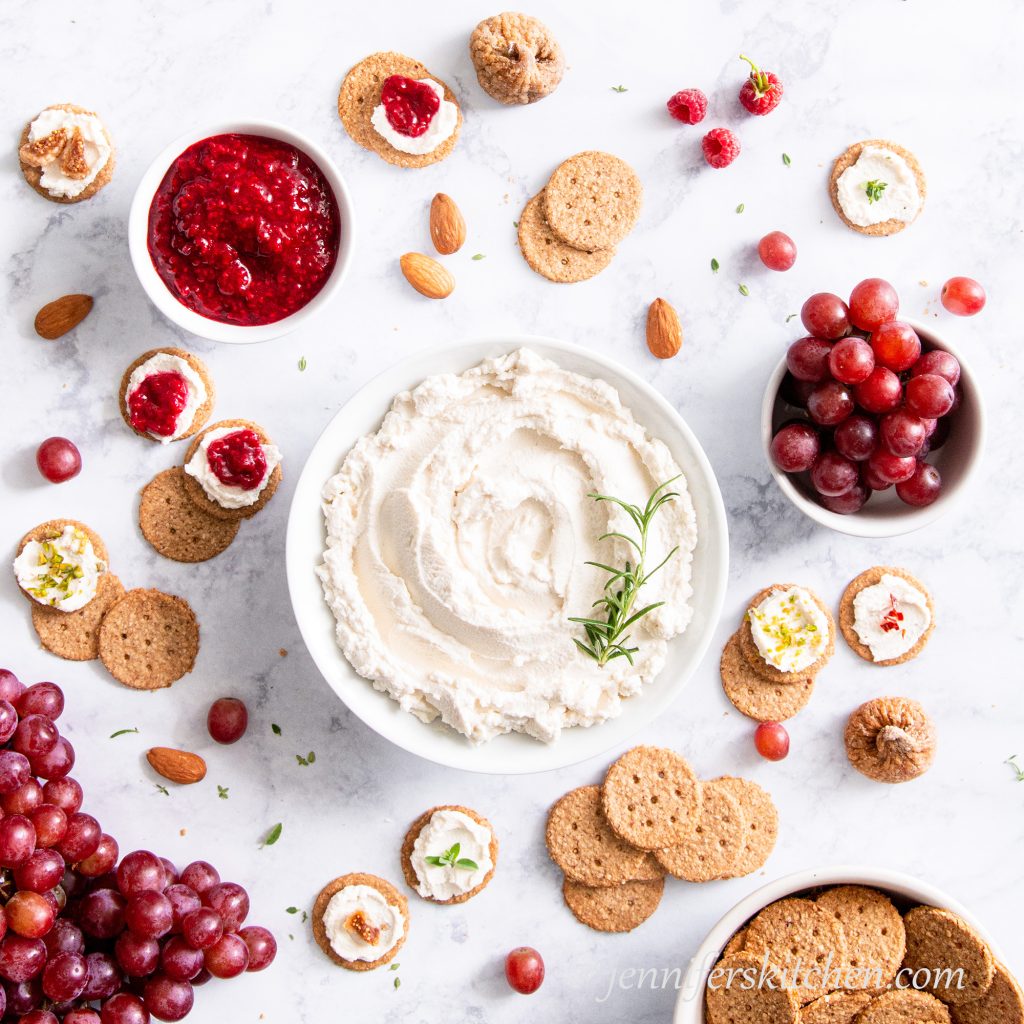 A bowl of vegan almond cheese with crackers, grapes, raspberry jam, and almonds.