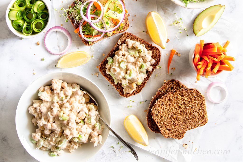 No-Mayo Vegan Chickpea Salad in a white bowl with open-face sandwiches topped with cucumber, sprouts, and onions.