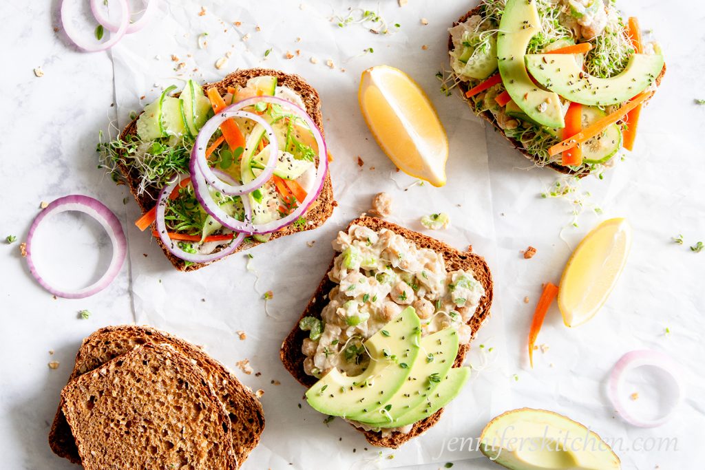 Open-face No-Mayo Vegan Chickpea Salad Sandwiches topped with avocado, cucumber, sprouts, and onions.