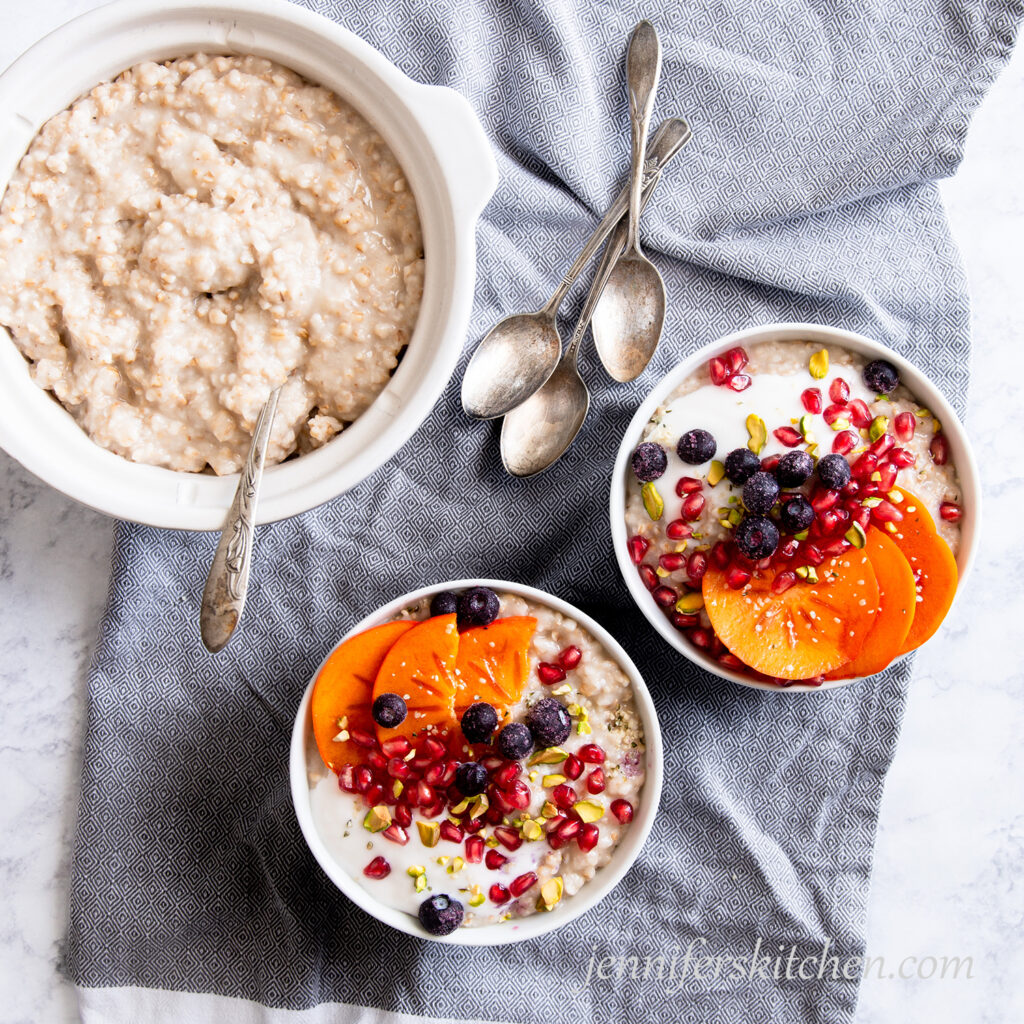 Oats in a crockpot/slow cooker with two bowls topped with blueberries, pomegranate, persimmon, and pistachios.