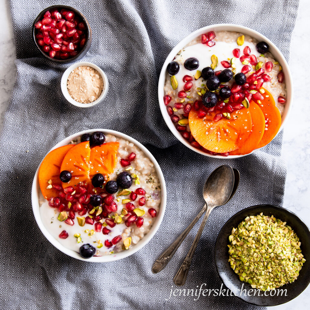 Two bowls of oats topped with blueberries, pomegranate, persimmon, and pistachios.