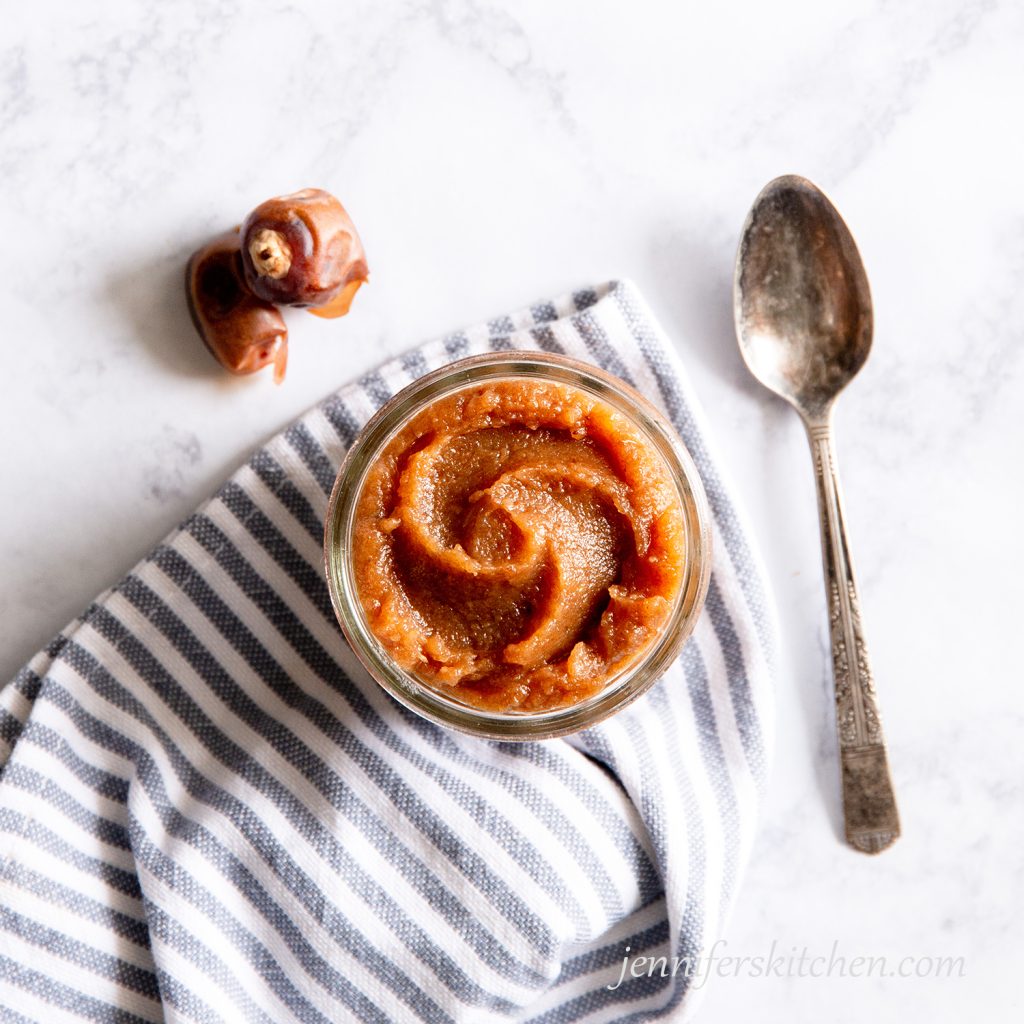 A jar of date paste on a towel with a spoon and a couple of dates.