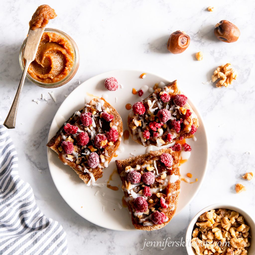 Toast spread with date paste and topped with raspberries and walnuts with a jar of date paste and a bowl of walnuts on the side.