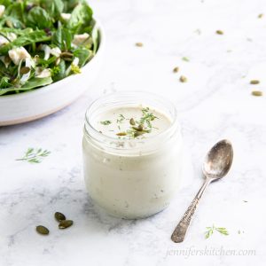 Side shot of Creamy Pumpkin Seed Dressing in a jar garnished with herbs and pumpkin seeds with a salad on the side.