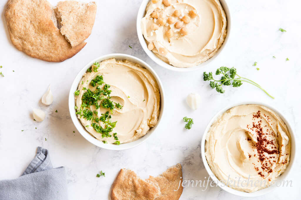 Three bowls of oil-free hummus topped with fresh parsley, garlic, and chickpeas with pita bread.