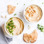 Two bowls of oil-free hummus topped with fresh parsley, garlic, and chickpeas.