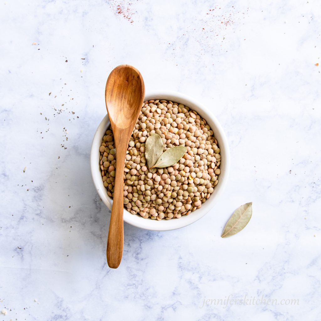 Uncooked brown lentils in a white bowl with bay leaves and a wooden spoon.