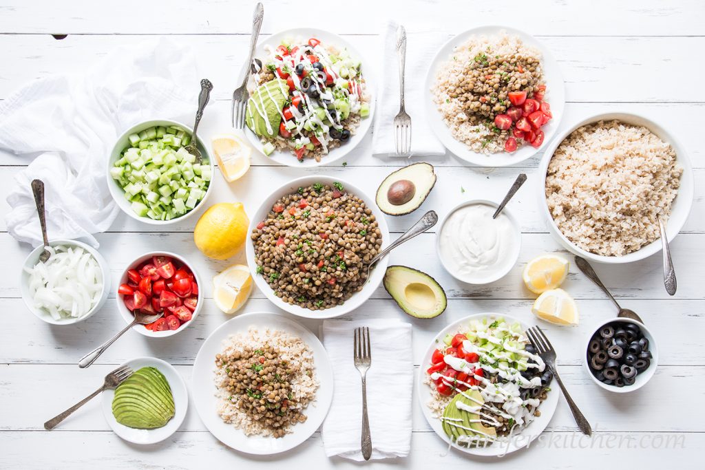 A meal of Mazidra with lentils, rice, cucumbers, tomatoes, vegan sour cream, sliced onions, avocado, olives, and lemon wedges.