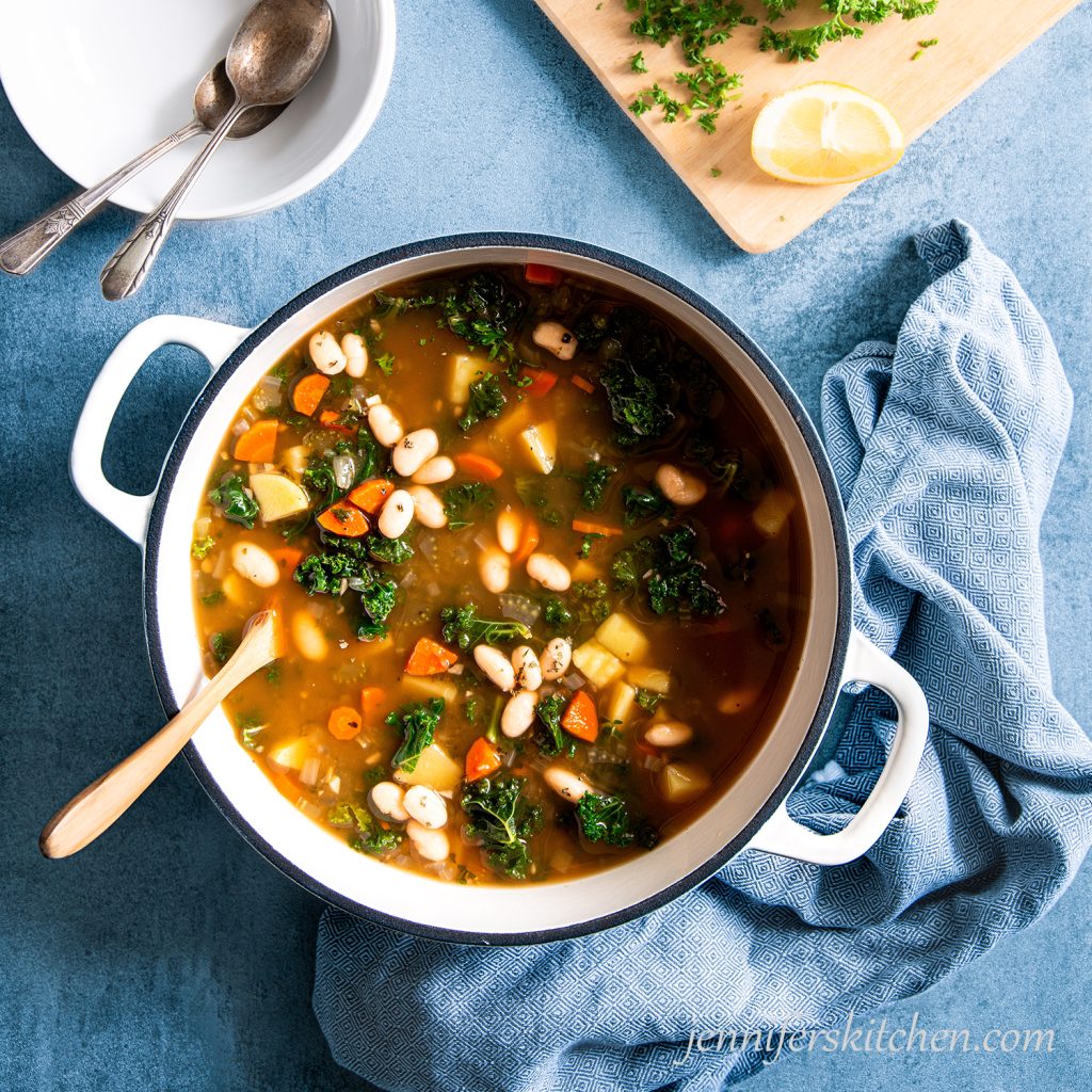 Bean Potato Kale Bean Soup in a soup pot on a blue background with a wooden spoon, a towel, and a stack of bowls.