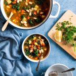 Bean Potato Kale Soup in a bowl on a blue background with a wooden spoon, a towel, and a soup pot.