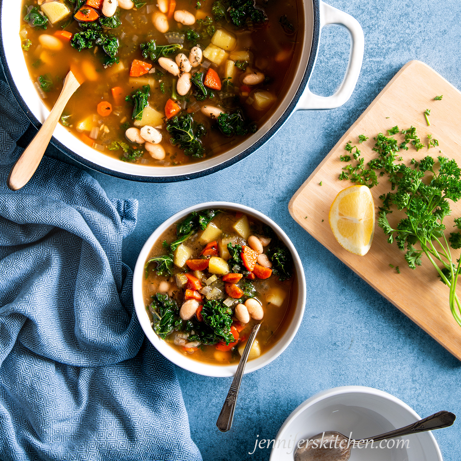 Bean Potato Kale Soup in a bowl on a blue background with a wooden spoon, a towel, and a soup pot.