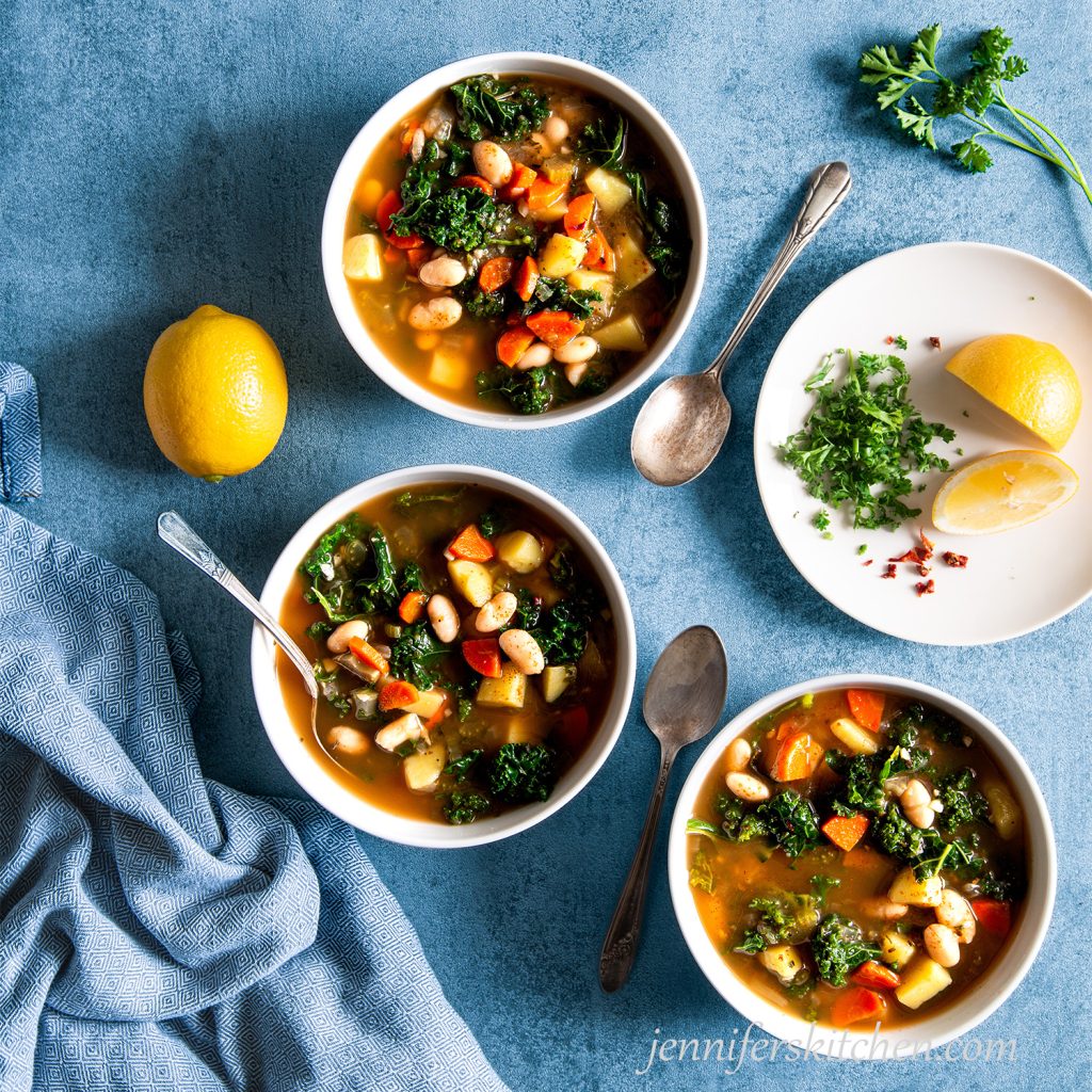 Three bowls of Potato Kale Bean Soup on a blue background with a towel.