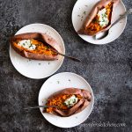 Three sweet potatoes on plates topped with vegan cashew sour cream and sprinkled with fresh herbs.