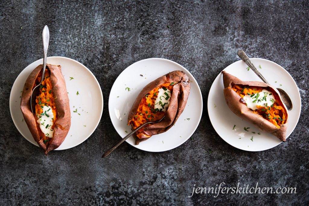 Three sweet potatoes on plates topped with vegan cashew sour cream and sprinkled with fresh herbs.